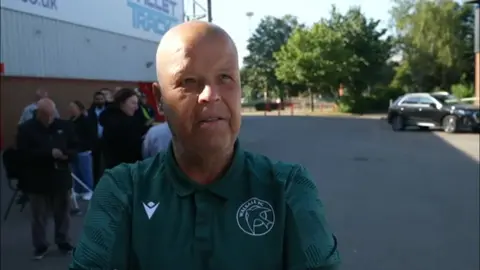 A man is wearing a green top including Walsall FC's club crest.