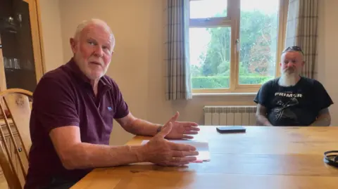 Two men sit at a wooden dining table. There is a document on the table and a phone. There is a window in the background showing greenery and trees.