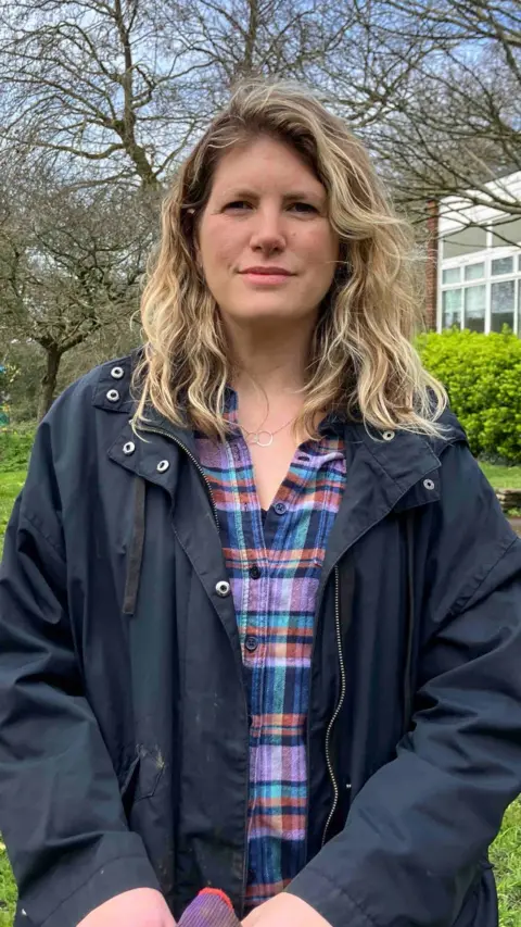 A woman with long blonde hair and a dark jacket and checked blue and red shirt stands in St Georges Park in Bristol