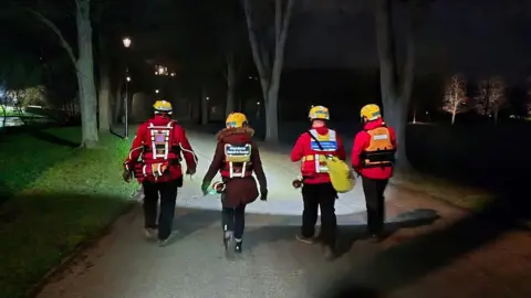 West Mercia Search and Rescue Four people pictured from the back walking in the dark. They are on a path and there are trees and grass on the path. They are wearing red jackets and yellow helmets. 