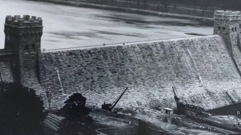 A black and white image of Derwent Dam, a large brick wall with two towers at either end. Cranes and trees can be seen at the bottom of the dam and there is a body of water to the left of it.