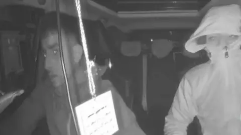 A grey screenshot image of CCTV footage, showing two people sat inside a car. The driver is looking forward, wearing a hooded jacket, with the passenger seen with a jacket zipped high over his face. A lanyard is hanging from the rearview mirror with writing on