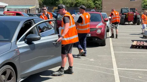 BBC A number of staff wearing high-vis orange jackets provide bottled water to people sitting in cars. 