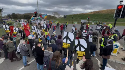 PA Media A group of protestors seen mostly from behind as they face a grassy bank. They stand in the road between traffic lights holding black and white CND signs saying Kick out Trump's Nukes and No War on Iran. 