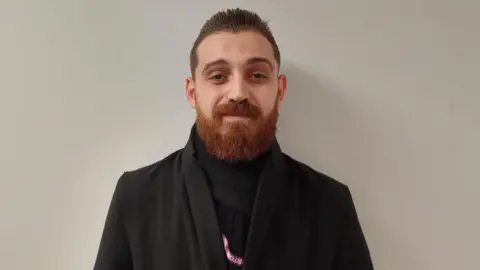 Mohammed is standing against a white background. He's wearing a black coat and scarf, and looking into the camera. He's a beard.