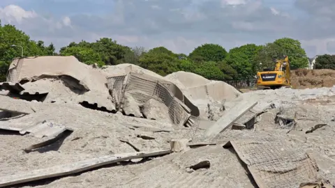 The site of a bomb detonation in Plymouth. A large crater has been left with debris strewn. A yellow digger is in the background.