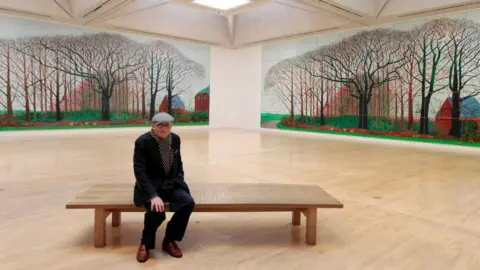 Getty Images British artist David Hockney poses for photographers next to his 2007 painting Bigger Trees Near Warter, which is displayed next to an identical photographic copy at the Tate gallery in London. He is wearing a dark suit and grey cap.
