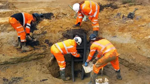Oxford Cotswold Archaeology Four archaeologists in hi-vis orange suits and black boots bending over a sandy soiled stepped site. In the middle is a blackened ladder with three rungs emerging from a watery pit.