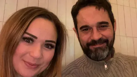 Family handout Kimberley Newark and Yann Trupiano pose for a selfie close to the camera with a blank wall behind them. Yann has a beard and is wearing a brown zip up jumper