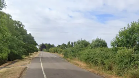Google A rural country road with fields on either side 