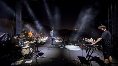 Roberto Panucci - Corbis/ Getty Images A band performing on stage. The photo is taken from behind the band, and in the distance you can see the audience. On the right, there is a man standing by a keyboard. On the left, there is a man sitting at a drum-set. In the middle, and further away, is a man standing, holding a microphone. All three are dressed all in black.