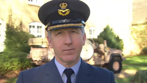 Group Captain Andy McIntyre is dressed in RAF uniform with a hast, blue suit, shirt and tie. Behind him out of focus is a building and a military vehicle.