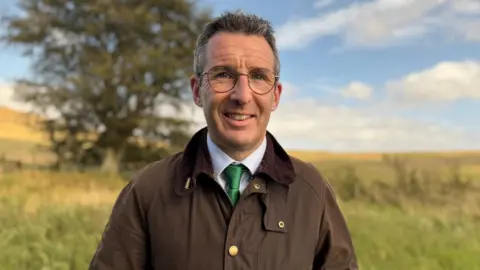 Andrew Muir is wearing a brown Barbour coat, blue shirt and a green tie. He has silver glasses on. Behind him is an open field with a single tree in it. He has short brown hair with some grey in it.