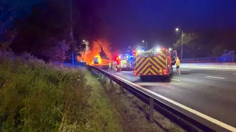 National Highways Two fire engines on the M15, with firefighters at the scene. There is a grass bank and trees to the left. Flames can be seen in the distance. 