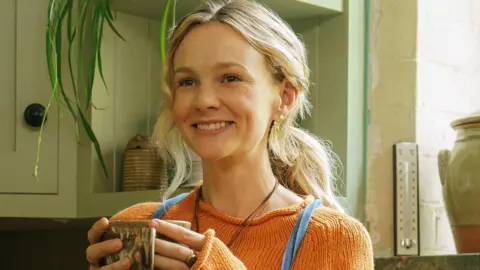NBCUniversal Mulligan has long blonde hair tied up in a low ponytail behind her back. She holds a mug in front of her with both hands. She wears an orange knitted jumper and you can see the thin straps of her blue dungarees. She is smiling, with kitchen cabinets and a window ledge behind her. 