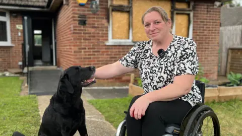 A woman with long brown hair in a wheel chair petting a black labrador, with a badly damaged home in the background