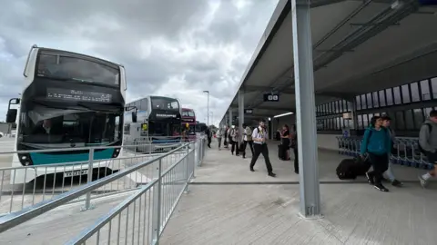 Buses lined up at a transport interchange, with passengers getting off and carrying suitcases