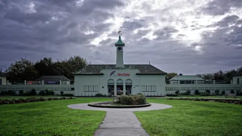 Laurence Cawley/BBC A front view of Peterborough Lido. It is a white art deco building with a painted sign showing a beach umbrella and the words "the lido". There is a lawned area to the front and a clock tower on top of the main building.