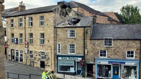 BBC A view of a street of tall, terraced, sandstone buildings. The two to the right are shops, with one of them, Bobs Barbers has a gaping hole in the tiled roof due to a collapsed chimney. Some rubble is strewn on the ground in front of the building and the street is taped off. 