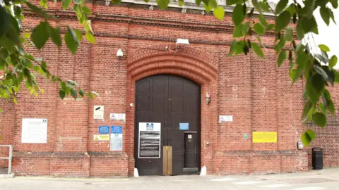 The large brown entrance door to HMP Norwich, which is in the middle of tall and wide brick walls. Branches of a leafy tree appear in the foreground of the image.