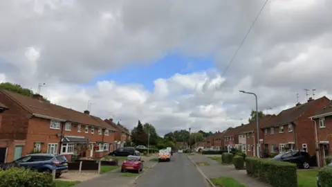A residential street lined with brick houses, parked cars, and small front gardens beneath a cloudy sky.