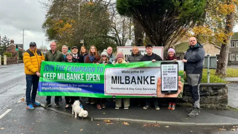 Adam Brierley Fourteen people including the MP for Fylde Andrew Snowden hold a sign saying 'Join the campaign to keep Milbanke open'. They are standing on the pavement outside the home. A small white dog is in front of the sign to the left.