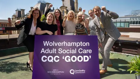 Seven people, six women and one man are standing on what looks to be a rooftop area of a building, around a large purple sign that reads "wolverhampton adult social care CQC good". They are cheering with their fists in the air in celebration