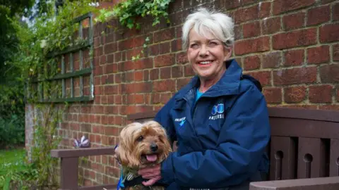 Battersea Ann Palmer, who has white, short hair, is white and has red lipstick, is sat on a bench with brown Yorkshire Terrier Jack