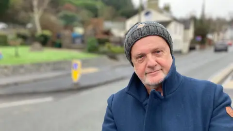 A man in an overcoat and beanie hat stood near a chicane created by a build-out in the road