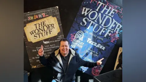 Middle aged man with a hoody on pointing behind him to The Wonder Stuff band posters