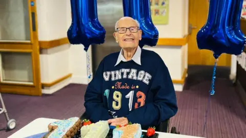 Cubbington Mill Care Home John Farringdon smiling at the camera in a blue 'vintage made in 1913' jumper, with three cakes spelling out '111' in front of him and blue balloons behind him