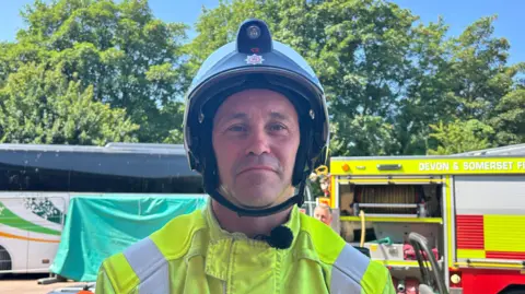 A firefighter with a yellow jacket and a dark coloured helmet. He has light stubble