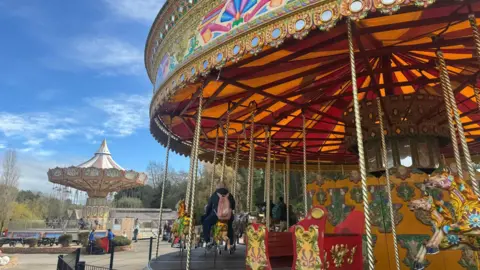 BBC/ Emily Johnson A brightly coloured fairground scene with an ornate carousel in the foreground, featuring painted horses and a red sleigh, and a larger swing‑style ride behind it, all set outdoors under a blue sky with scattered clouds.