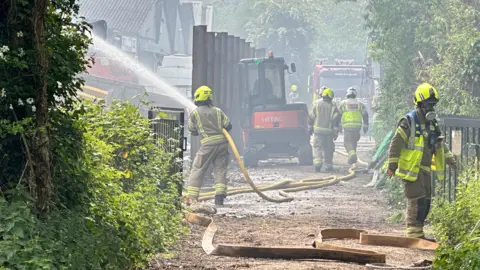 Eric Johnson/BBC A close up of firefighters dousing the flames at the recycling centre. A fire engine is parked up at the end of the lane. 