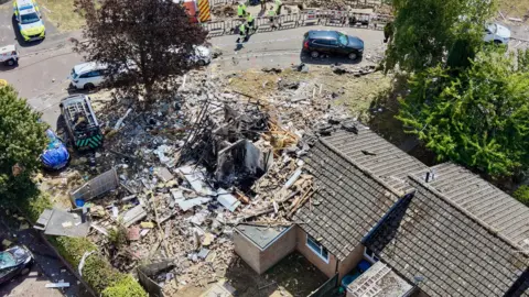 Ant Saddington/BBC An aerial shot of three bungalows. The one on the far left is completely destroyed with just four internal walls remaining. There is debris everywhere. There is a police car parked nearby and people in high-vis vests.