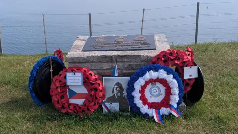 Gary Hadden/@garyh_creates A memorial for Czech pilot Alois Vasatko. There is a picture of him surrounded by poppy wreaths and tributes. The memorial is made from stone and has a plaque on the top of it. It is placed on a grass cliff edge with the sea visible in the background.