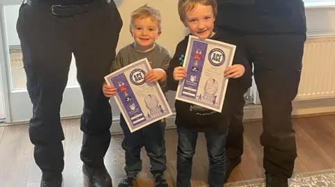Two male police officers in their uniforms, standing next to each other facing the camera, you can only see their legs as they are much taller than the children. In front of them are two little boys, aged three and five, the boy on the left had blonde hair, the boy on the right had brown hair. They are holding police certificates. 