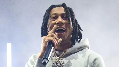 Digga D wearing his hair in braids, a light grey hoodie with a huge necklace and a pendant spelt BMR dangling from the chain. He is looking happy as he sings into a wireless mic he is holding with his right hand.