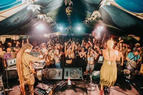 Recompose A view from behind an act performing on stage to a crow of people dancing in a large marquee
