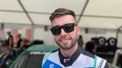 A man in sunglasses and a racing suit smiling