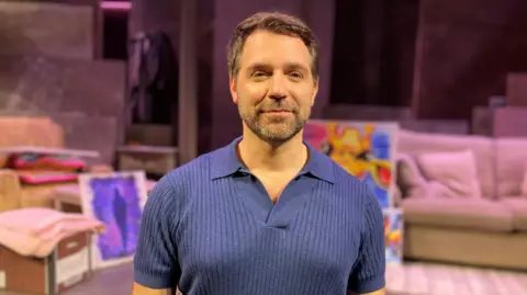 A man with brown hair and stubble wearing a blue polo t-shirt.