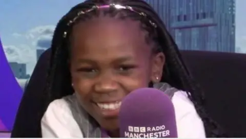 Young Elphaba smiles while sat in front of a microphone in a BBC studio 