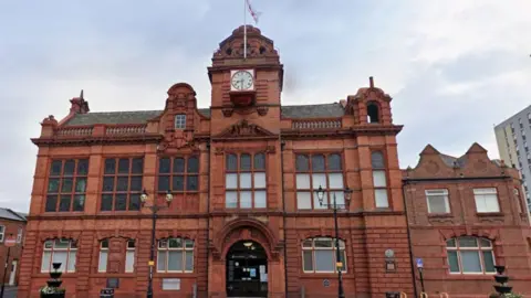 Google Jarrow Town Hall. An ornate, deep red brick building with large leaded windows on the first floor. There is a central dome with a clock and flagpole.