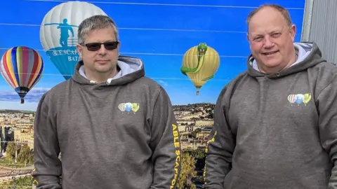 Adrian Connor Two men in grey hooded sweatshirts stand in front of an image of hot-air balloons flying over the city of Bath