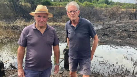 Henry Godfrey-Evans/BBC Two men in dark clothing looking at the camera with a small stream and a burnt landscape behind them