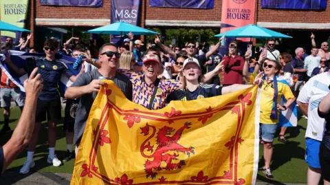 Scotland fans holding a beer in one hand and the Scottish flag in another basking in the sunshine of a beer garden at a Scottish fan park in Boston