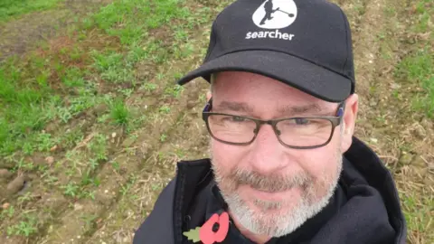 Malcolm Weale Malcolm Weale, in a field, smiling at the camera in what appears to be a selfie taken holding his arm above and in front of his face. He is wearing a black baseball cap, black top, and a red poppy pin. He has glasses and a grey and brown beard. There is grass in the field behind him. 