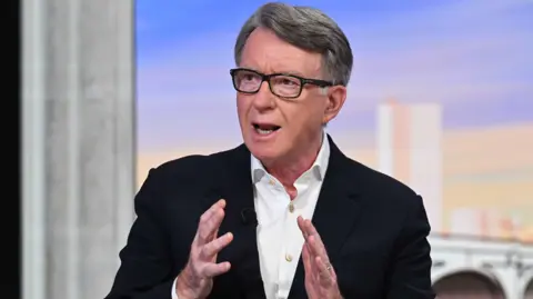 Close-up of Peter Mandelson, wearing a black jacket and white button-up shirt and black spectacles, speaking at a BBC studio.