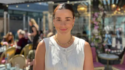 Zara Kramer standing at an outdoor café with a pergola roof and string lights, surrounded by tables, chairs, and floral decorations. Other patrons are visible in the background.
