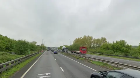 Google A Google view of a dual-carriageway, with multiple cars and lorries. On both sides of the road are trees and street lights.